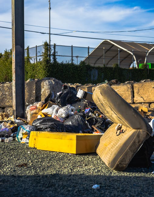 An outdoor scene showing a large pile of mixed household rubbish and waste materials scattered on a gravel surface beside a low stone wall. The debris includes black plastic bin bags, a yellow plastic storage container, and a discarded old sofa cushion with beige fabric and visible stuffing, all positioned towards the right side of the image. In the background, there is a metal utility pole and overhead power lines, with a green hedge and a partially covered sports court or tennis enclosure visible beyond. The sky is clear with some clouds, and the lighting indicates it is daytime with ample natural sunlight illuminating the scene. The image suggests the cleanup or removal of unmanaged waste, which a private waste disposal service such as Rubbish Clearance Hackney might take on as part of their rubbish or waste management services, particularly in contexts where alternative disposal options are sought to avoid hidden charges or to handle bulky items and household refuse on-site.