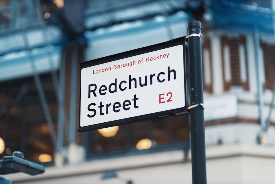 A close-up view of a white rectangular street sign mounted on a black pole, indicating 'Redchurch Street' in the London Borough of Hackney, with the postcode 'E2' in red. The sign features black text with a small red line and the words 'London Borough of Hackney' printed above the main name. In the background, there is a modern building with large glass windows reflecting ambient light, and scaffolding structures along the upper facade, suggesting ongoing construction or renovation. The scene is illuminated by natural daylight, creating a clear and neutral environment that subtly emphasizes urban infrastructure. This setting is relevant to rubbish removal services, such as those provided by Rubbish Clearance Hackney, by illustrating an exterior city location where waste management and private clearance might be necessary for maintaining street cleanliness and proper waste disposal outside residential or commercial premises.