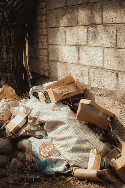 An outdoor scene showing a large pile of mixed household rubbish and waste materials scattered on a gravel surface beside a low stone wall. The debris includes black plastic bin bags, a yellow plastic storage container, and a discarded old sofa cushion with beige fabric and visible stuffing, all positioned towards the right side of the image. In the background, there is a metal utility pole and overhead power lines, with a green hedge and a partially covered sports court or tennis enclosure visible beyond. The sky is clear with some clouds, and the lighting indicates it is daytime with ample natural sunlight illuminating the scene. The image suggests the cleanup or removal of unmanaged waste, which a private waste disposal service such as Rubbish Clearance Hackney might take on as part of their rubbish or waste management services, particularly in contexts where alternative disposal options are sought to avoid hidden charges or to handle bulky items and household refuse on-site.