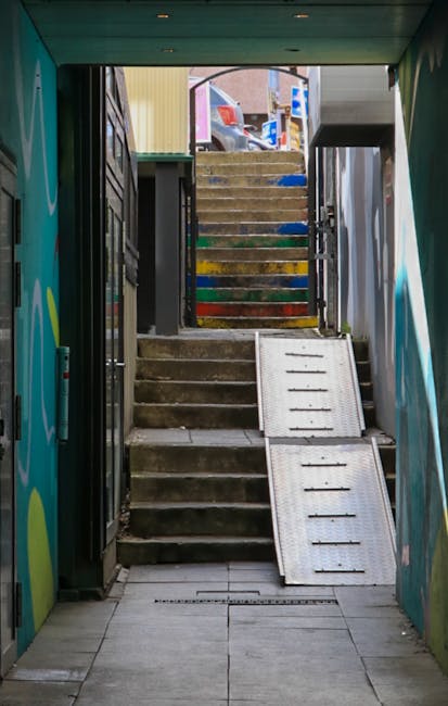 The image depicts a narrow outdoor staircase viewed from inside a building or underpass, with concrete steps leading upward to a street level. The staircase is flanked by metal safety railings on both sides, and at the top, there are some colorful wooden steps painted in various shades including yellow, green, blue, and red, which appear to be worn and weathered. A metal ramp with a textured surface is positioned to the right of the concrete steps, potentially used for wheelchair access or trolley movement. The environment outside the stairway shows parked vehicles, including a white van and a red car, and some urban elements like buildings and signage are visible in the background. The pathway inside leads to the street level, suggesting this could be an alternate access point for waste or rubbish collection, often associated with private waste handling or on-site clearance services such as those offered by Rubbish Clearance Hackney. The lighting suggests daytime, with natural light illuminating the scene.
