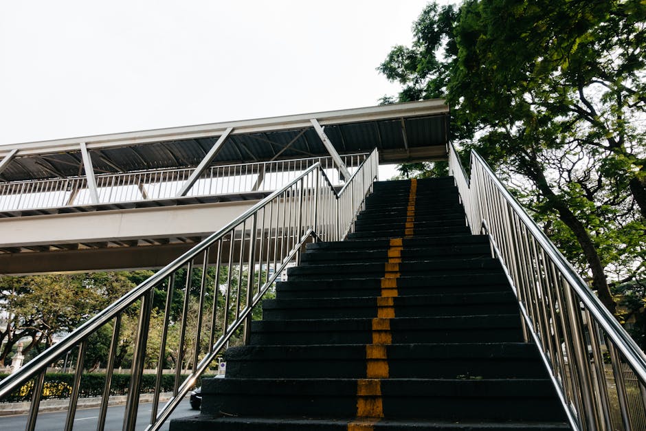 The image depicts a narrow outdoor staircase viewed from inside a building or underpass, with concrete steps leading upward to a street level. The staircase is flanked by metal safety railings on both sides, and at the top, there are some colorful wooden steps painted in various shades including yellow, green, blue, and red, which appear to be worn and weathered. A metal ramp with a textured surface is positioned to the right of the concrete steps, potentially used for wheelchair access or trolley movement. The environment outside the stairway shows parked vehicles, including a white van and a red car, and some urban elements like buildings and signage are visible in the background. The pathway inside leads to the street level, suggesting this could be an alternate access point for waste or rubbish collection, often associated with private waste handling or on-site clearance services such as those offered by Rubbish Clearance Hackney. The lighting suggests daytime, with natural light illuminating the scene.