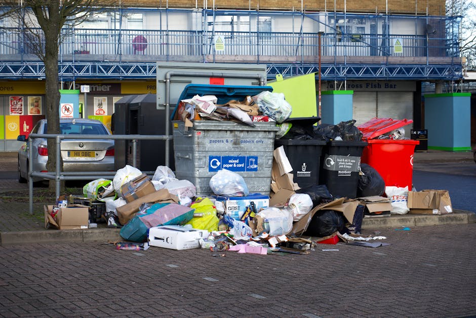 A large pile of mixed domestic waste and recyclable materials is situated on a paved urban street corner, with various black and black-and-white bin bags, flattened cardboard boxes, and plastic bags overflowing from a central grey recycling bin labeled for mixed paper and cardboard. Surrounding the pile are additional waste containers including a red bin, black bins, and a black recycling box, all positioned on the pavement next to a metal railing. Behind the waste scene, there is a commercial building with a blue scaffolding structure, and storefront signs indicating businesses, including one with a yellow storefront. A parked silver car is visible to the left of the waste collection area. The scene is set outdoors during daytime with overcast lighting, and a leafless tree is partially visible to the left, indicating a winter or late autumn setting. This image illustrates an instance of private rubbish accumulation outside an urban retail area, highlighting the need for waste disposal services such as those provided by Rubbish Clearance Hackney, which offers alternative rubbish removal options outside municipal collection.