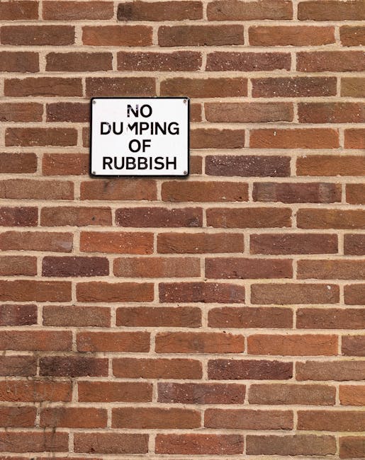 A rectangular metal sign with a white background and black text, mounted on a brick wall. The sign reads 'NO DUMPING OF RUBBISH' with the words 'DUMPING' and 'RUBBISH' separated onto different lines, and the word 'OF' in smaller font. The wall behind the sign is constructed of reddish-brown bricks with visible mortar joints, arranged in a horizontal running bond pattern. The bricks have a slightly textured surface with some variation in color tones, ranging from deep red to lighter shades of orange and brown. The lighting creates a faint shadow of the sign on the brick wall, emphasizing the three-dimensional aspect of the sign and the textured surface of the bricks. This scene is typical of urban areas where waste management and private rubbish removal services, such as those provided by Rubbish Clearance Hackney, are often employed to manage refuse collected from rental properties or estate cleanups.