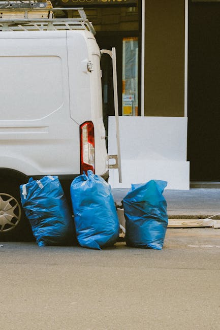 The image shows the rear side of a white delivery van parked on a city street, with three large blue rubbish bags placed on the pavement beside it. The bags are made of thick plastic with slight creases and are filled with mixed rubbish, standing upright on their bases. The van's rear door is closed, and part of the vehicle's taillight and wheel are visible in the foreground. Behind the van, there is a building with large glass windows and a door, suggesting a commercial or retail environment. The pavement is smooth, and the scene is lit by natural daylight, creating a neutral, straightforward view of private rubbish collection. Occasional details such as a yellow sticker on the van's door and a small window further indicate an urban setting where independent waste disposal methods like on-site clearance or collection by waste removal services might be employed, aligning with the context of private rubbish removal services provided by companies like Rubbish Clearance Hackney.