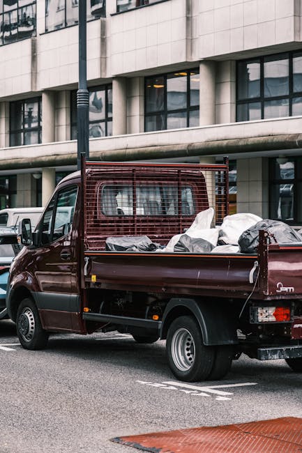 A white panel van parked on a city street with its rear doors open, revealing a large accumulation of waste materials and rubbish bags inside. The back of the van is filled with various debris, including cardboard boxes, plastic bags, and loose rubbish, some of which is stacked on the vehicle's roof. Several black rubbish bags are placed on the ground next to the van, with some leaning against the vehicle and others nearby on the pavement. A small hand truck with a black bag and a few scattered items is positioned in front of the open doors. The van is situated next to a multi-storey building with a light-colored facade, featuring narrow windows and architectural details typical of urban architecture. The environment appears to be an alley or a service street, with no other vehicles or pedestrians visible in the scene. This image underscores the type of waste transfer or on-site rubbish clearance services that companies like Rubbish Clearance Hackney might provide as an alternative to public waste collection, suitable for private rubbish disposal in urban environments.