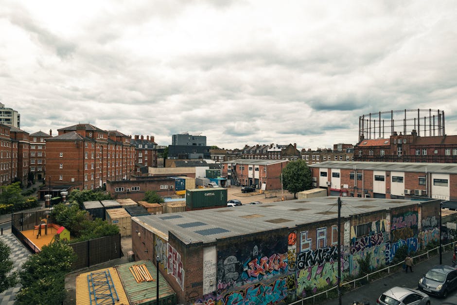 A daytime urban scene reveals a variety of low-rise brick buildings and industrial structures situated behind a fenced area with graffiti-covered walls. In the foreground, there is a flat-roofed warehouse or storage unit with a weathered appearance, featuring a mix of metallic and concrete textures. To the left, a small children's playground with a brightly colored slide and protective flooring is visible, bordered by trees and shrubs that add patches of green amidst the concrete environment. Several shipping containers, including a green one marked 'CAPITAL', are scattered around the area, indicating logistics or storage activity. The background showcases multiple residential and commercial buildings with brick facades, along with a circular metal framework structure, possibly an industrial tank or silos, set against a cloudy sky with diffused lighting. This scene exemplifies a typical mixed-use urban landscape associated with private waste handling and rubbish removal services, such as those provided by Rubbish Clearance Hackney, highlighting the environment where on-site clearance or alternative waste disposal methods might occur.