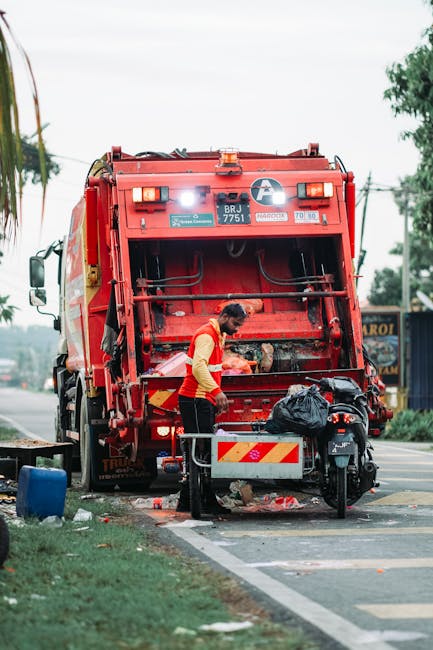 A worker in a high-visibility yellow and red vest is operating a red rubbish collection truck parked on the side of a road in Hackney. The truck’s rear compactor area is open, revealing mechanical components and a partially visible hydraulic system. The worker is leaning slightly forward, handling a black garbage bag that is placed on a small platform attached to the vehicle. In the foreground, a small motorbike is parked close to the curb, with a black plastic bag resting on its seat. The scene is set on a paved street with a grassy verge to the left, and a blue bin or container is visible on the grass nearby. The background includes some trees, a few utility poles with electrical wires, and distant building structures, indicating an urban environment. The overcast sky creates diffuse lighting, and the overall scene emphasizes on-site waste collection activity typical of private rubbish removal services, such as those provided by Rubbish Clearance Hackney, aligning with alternative waste handling methods outside local authority services.