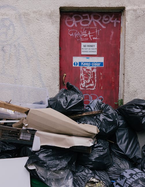 A pile of black plastic rubbish bags stacked against a rough, beige exterior wall with visible texture and small patches of discoloration. Some bags are partially torn or bulging, indicating filled waste, and they are placed in front of a red metal door covered with graffiti tags in white and pink. The door has various stickers attached, including a white one with the message 'NO MONEY, KEEP OFF' and a blue sticker with the phrase 'Keep clear.' There are also some loose cardboard and wood pieces, along with other miscellaneous waste materials visible among the bags. The scene appears to be outdoors on a pavement or alleyway, with natural light evenly illuminating the cluttered area, suggesting a scenario of on-site waste collection or private rubbish disposal, as handled by services like Rubbish Clearance Hackney.