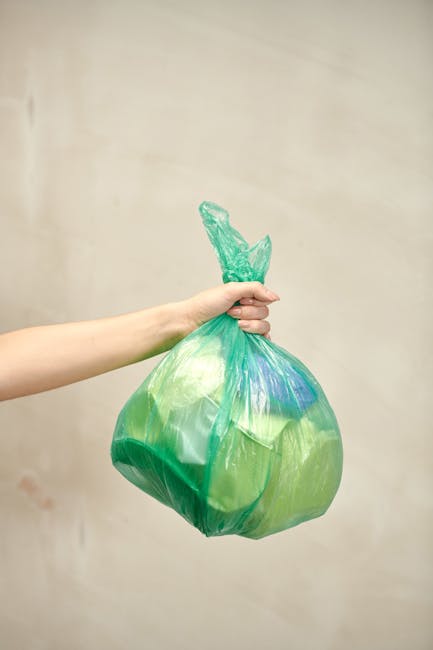 A person with fair skin is holding a green plastic rubbish bag by the tied top with their right hand, extending it outward against a plain, light beige wall. The bag appears to be filled with waste or discarded materials and has a slightly crumpled texture with visible folds and creases, reflecting light softly. The tied knot at the top of the bag is twisted and protrudes slightly, maintaining the bag's sealed shape. The person's arm, visible from the wrist to forearm, is positioned on the left side of the image. The background is plain and neutral, emphasizing the activity of handling waste, which pertains to rubbish disposal and waste management services. This scene could reflect the process of preparing household waste for private collection or on-site clearance, as offered by waste removal companies like Rubbish Clearance Hackney.
